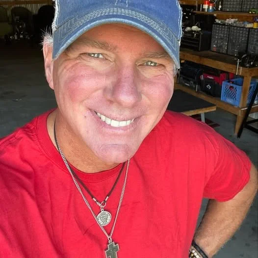A man wearing a blue baseball cap, red shirt, and necklaces smiling at the camera inside a workshop with shelves and tools in the background.