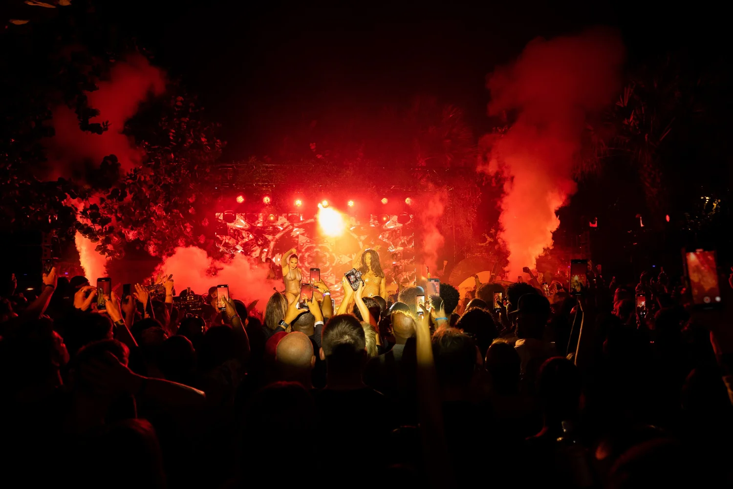 Nighttime outdoor concert with a crowd, stage, and performers, red lighting, smoke effects, and people taking photos.