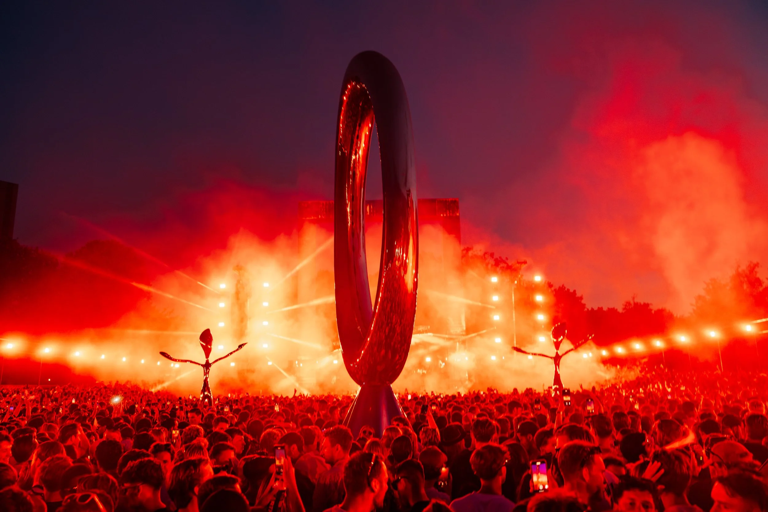 A large crowd of people at an outdoor event with bright orange lights, smoke, and fireworks in the background. There are two robot-like sculptures with outstretched arms among the crowd, and a large metallic ring structure in the center.