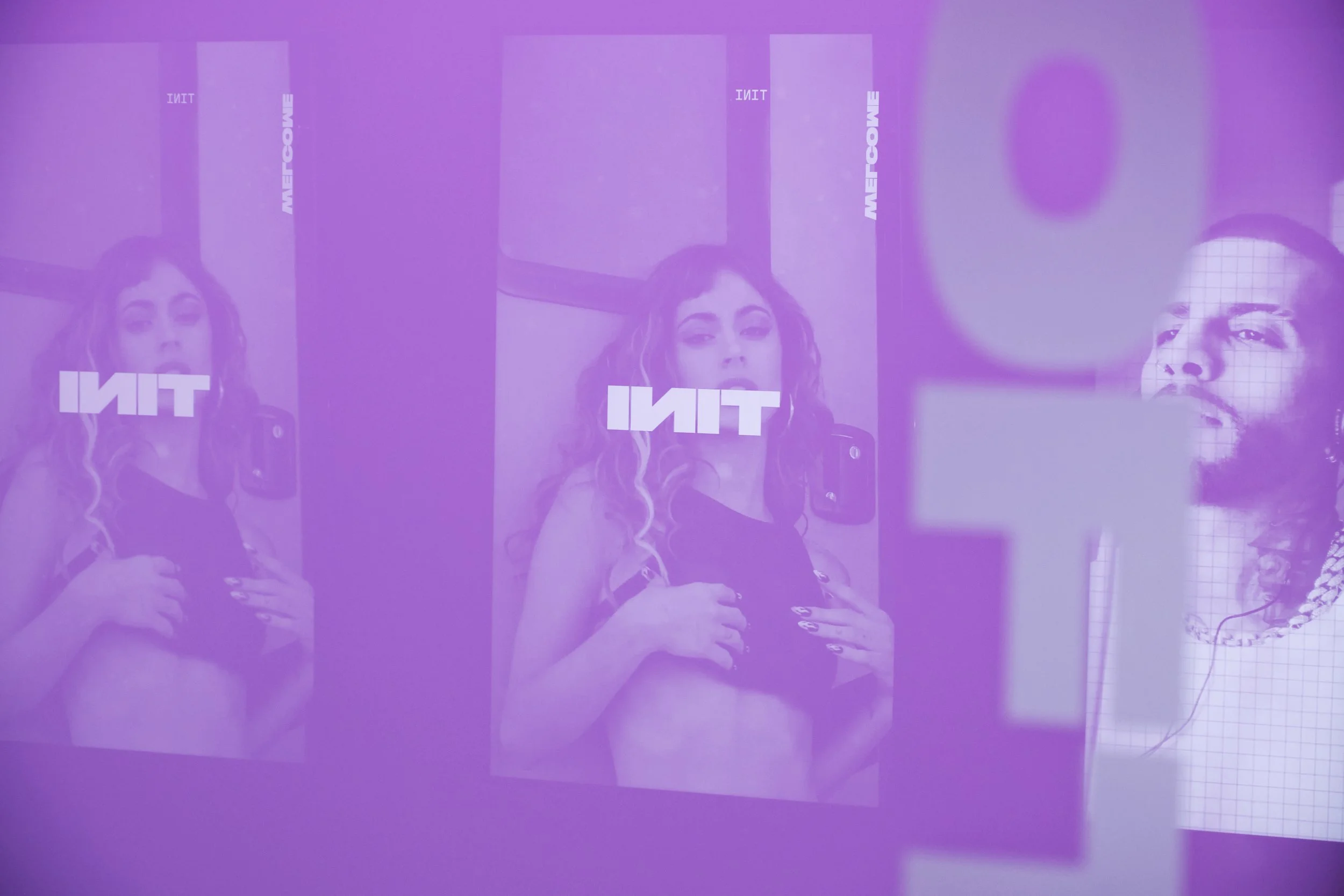 A wall with multiple mirrors or screens showing a woman in a black top with detailed nails, posing with her hands on her chest. The images are overlaid with a purple hue and the word 'TINI' appears multiple times.