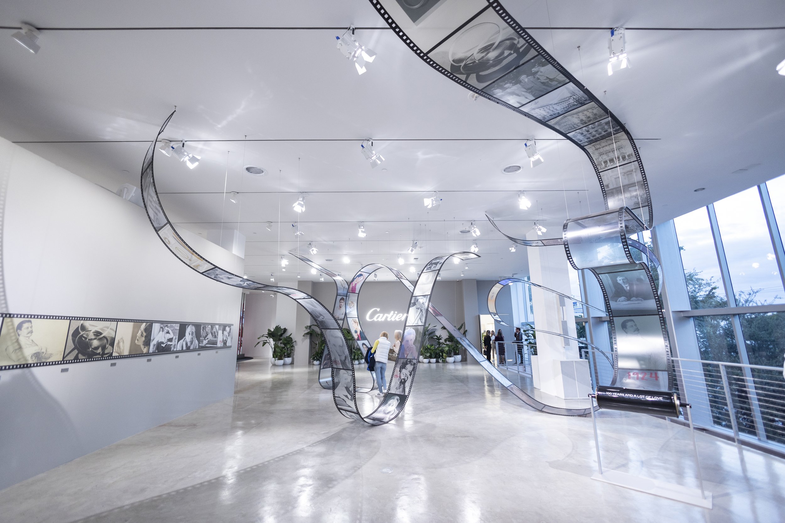 Museum interior with a modern art installation of film strip sculptures, black and white photographs on the wall, and large windows with a city view.