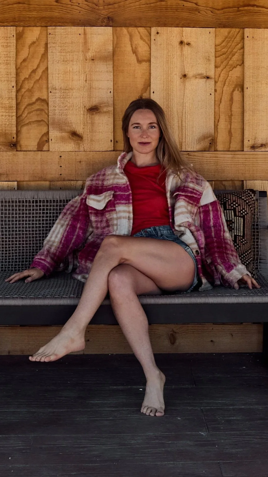 A woman sitting on a cushioned outdoor bench with crossed legs, wearing a red top, denim shorts, and a plaid jacket, against a wooden wall.
