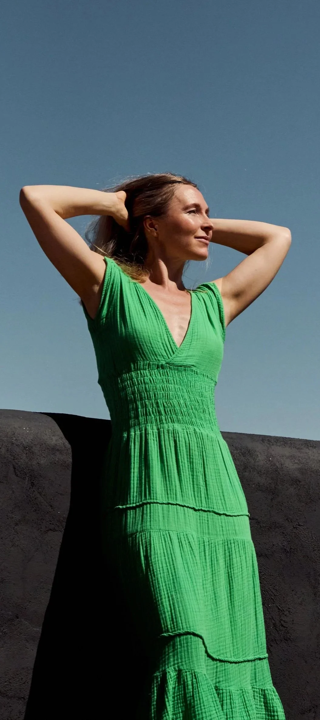 A woman in a bright green dress outdoors against a clear blue sky with her hands behind her head, facing slightly to the right.