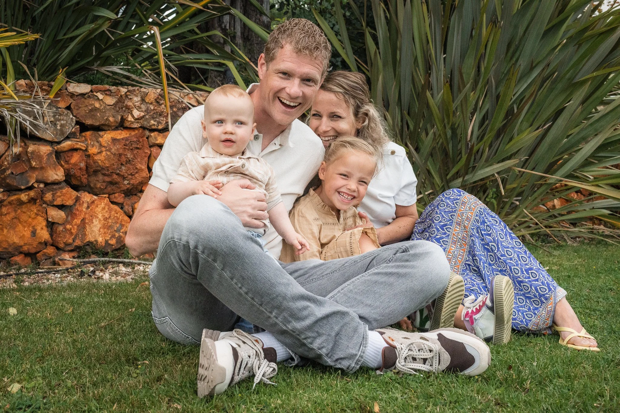 A smiling family of five sitting outdoors on grass, with large green plants and rocks in the background. The dad, mom, two young daughters, and a young toddler are happily posing.