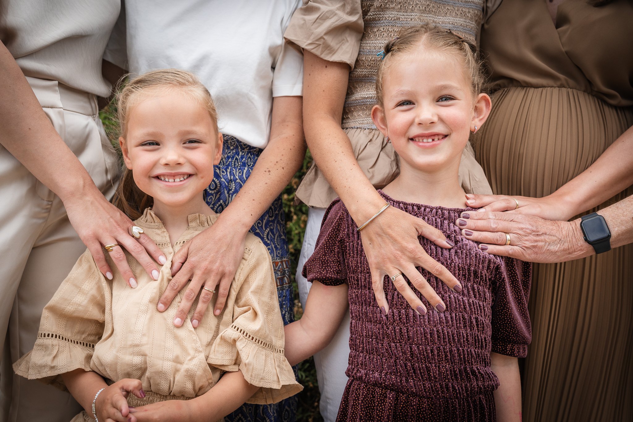 Two young girls and several adult women, partially visible, standing close together outdoors, with their hands on their chests, smiling.