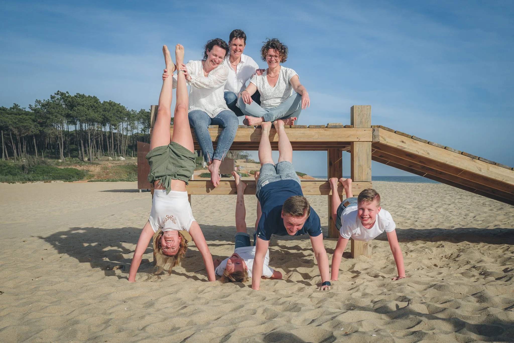 Group of children and adults forming a human pyramid on a sandy beach under a blue sky, with a wooden structure in the background.