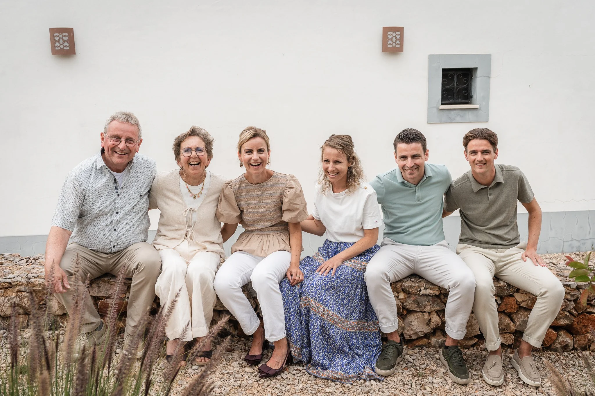 A group of six people, three men and three women, sitting together outdoors against a white wall, smiling and posing for the photo.