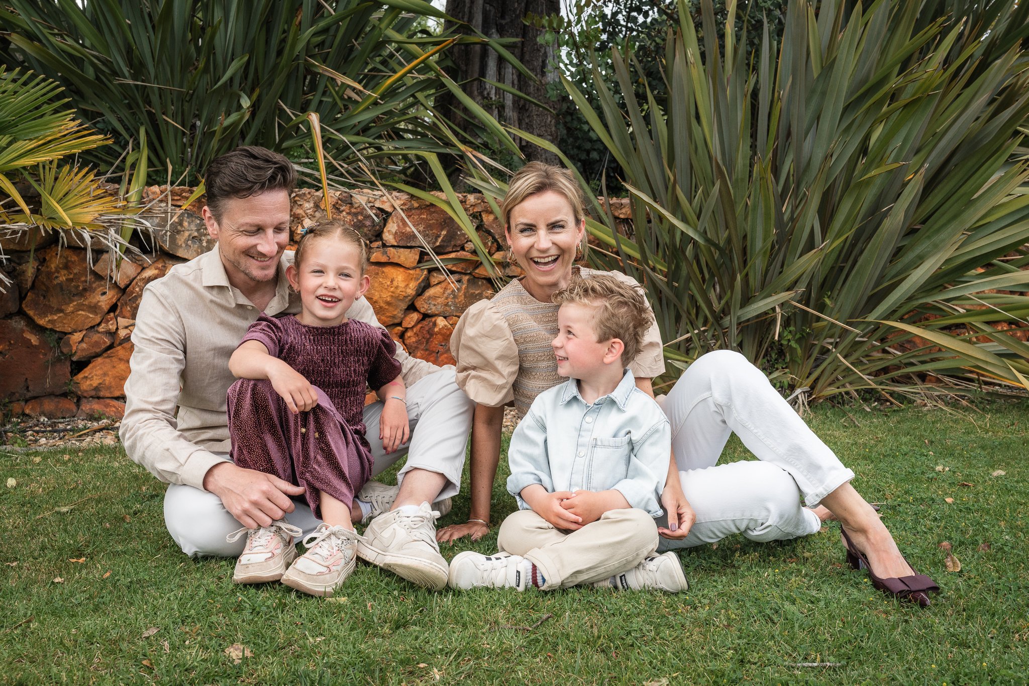 A happy family sitting on grass in front of large green plants and a stone wall, including two adults and two children, all smiling and enjoying each other's company.