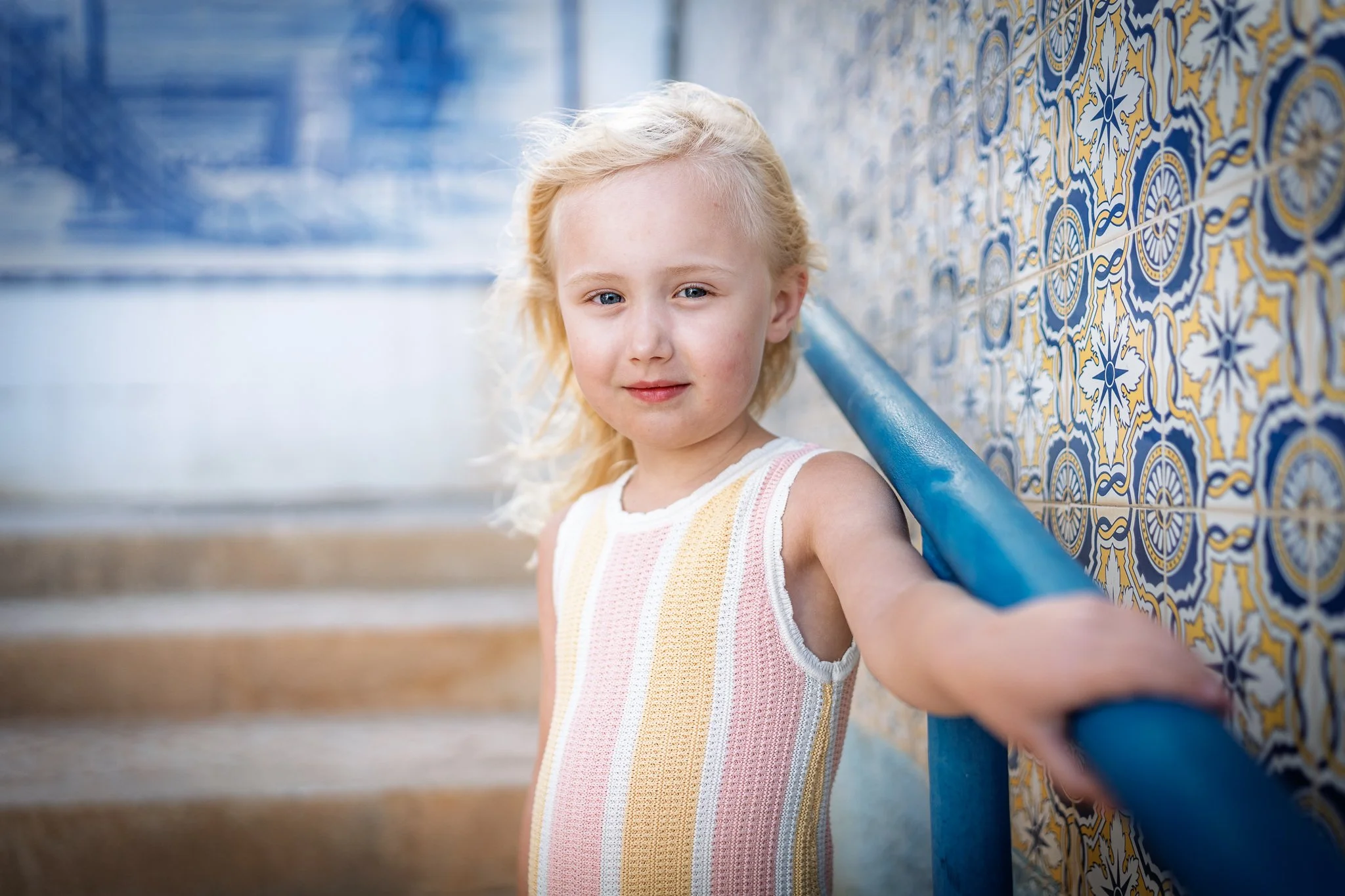 Young girl with blonde hair and blue eyes taking a selfie near a colorful patterned wall outside.