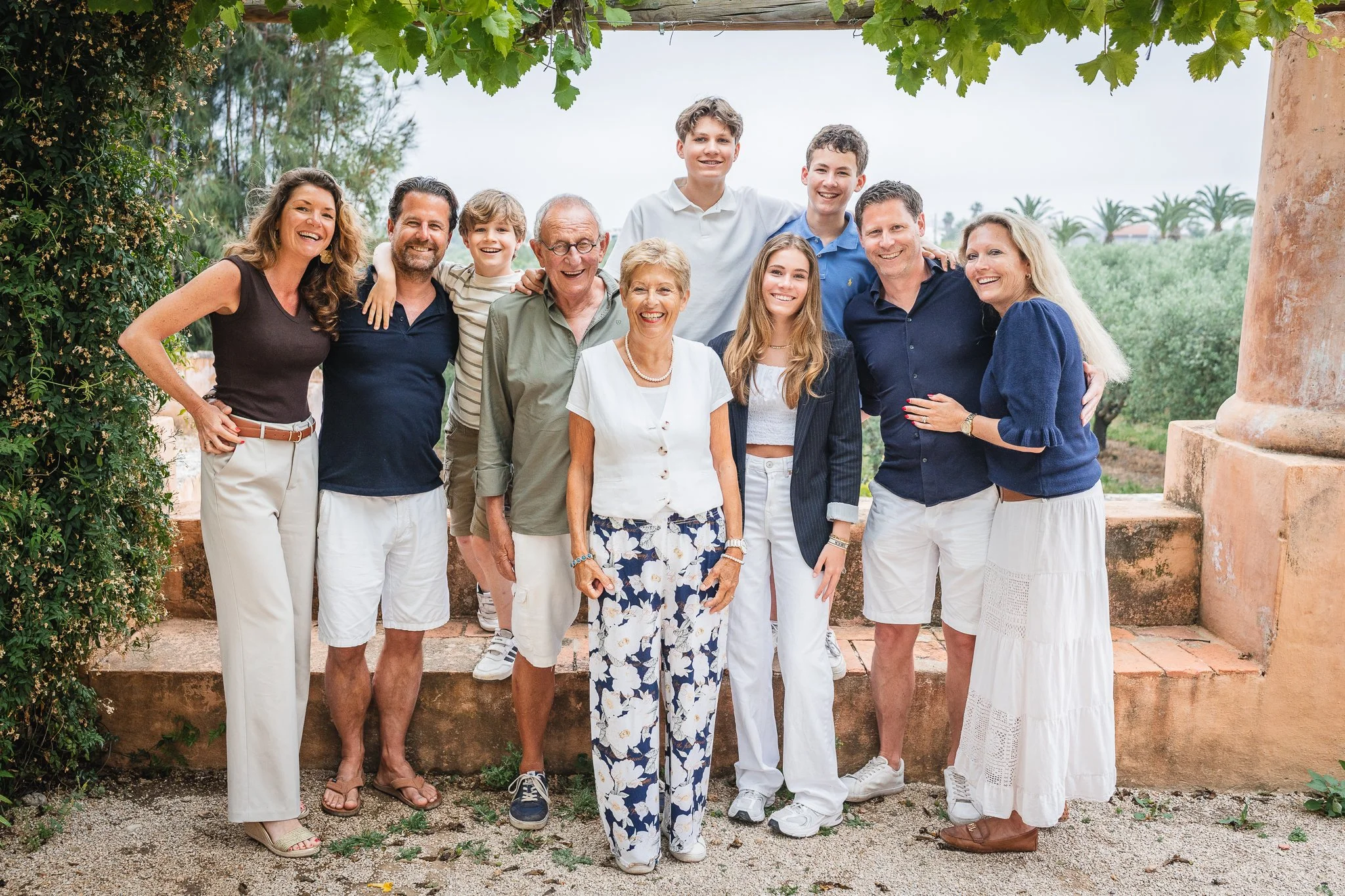 A large group of people, including children and adults, standing outdoors under a canopy of green leaves, smiling for a photo.