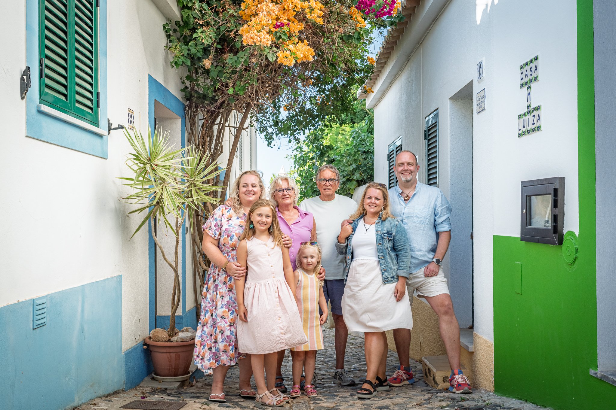A group of seven people, diverse in age and gender, standing together in a narrow street with colorful buildings and tropical plants. The group is smiling and enjoying a sunny day.