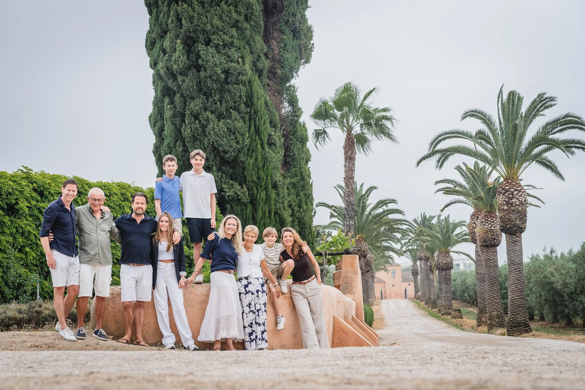 Group of people posing outdoors among palm trees and lush greenery, with some buildings visible in the background.