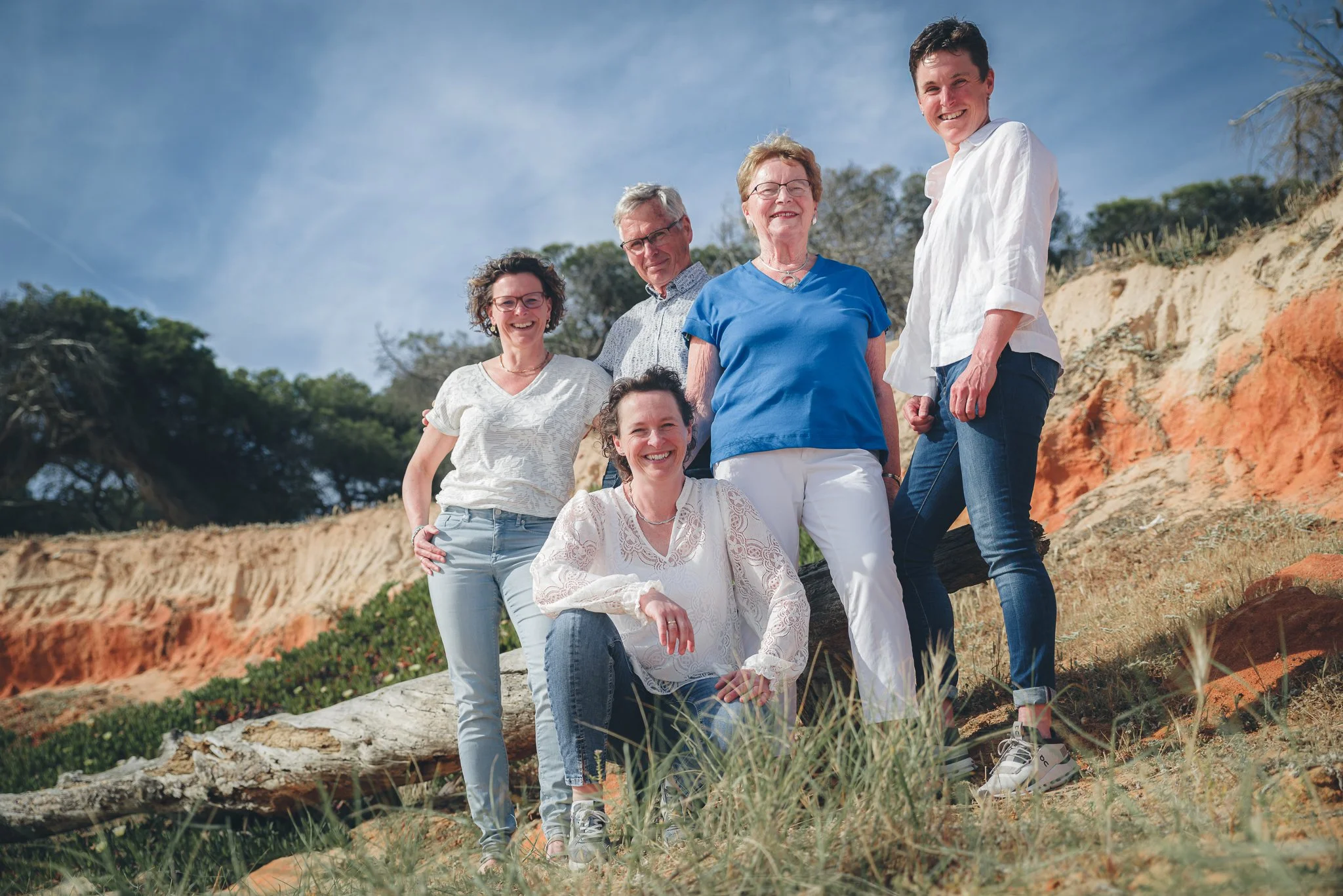 Group of six people, four women and two men, standing outdoors on a rocky terrain with trees and blue sky in the background, smiling and posing for the photo.