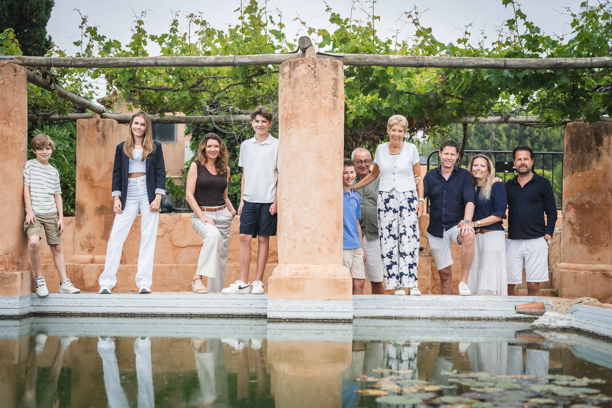 A group of eleven people, including children and adults, standing outdoors on a stone structure with a pond in the foreground, green grapevines and trees in the background, smiling and posing for the photo.