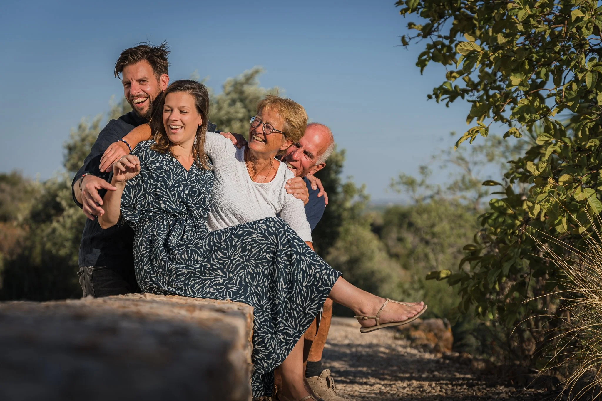 Four people are sitting on a log outdoors, enjoying each other's company and smiling. They are surrounded by trees and a clear blue sky.