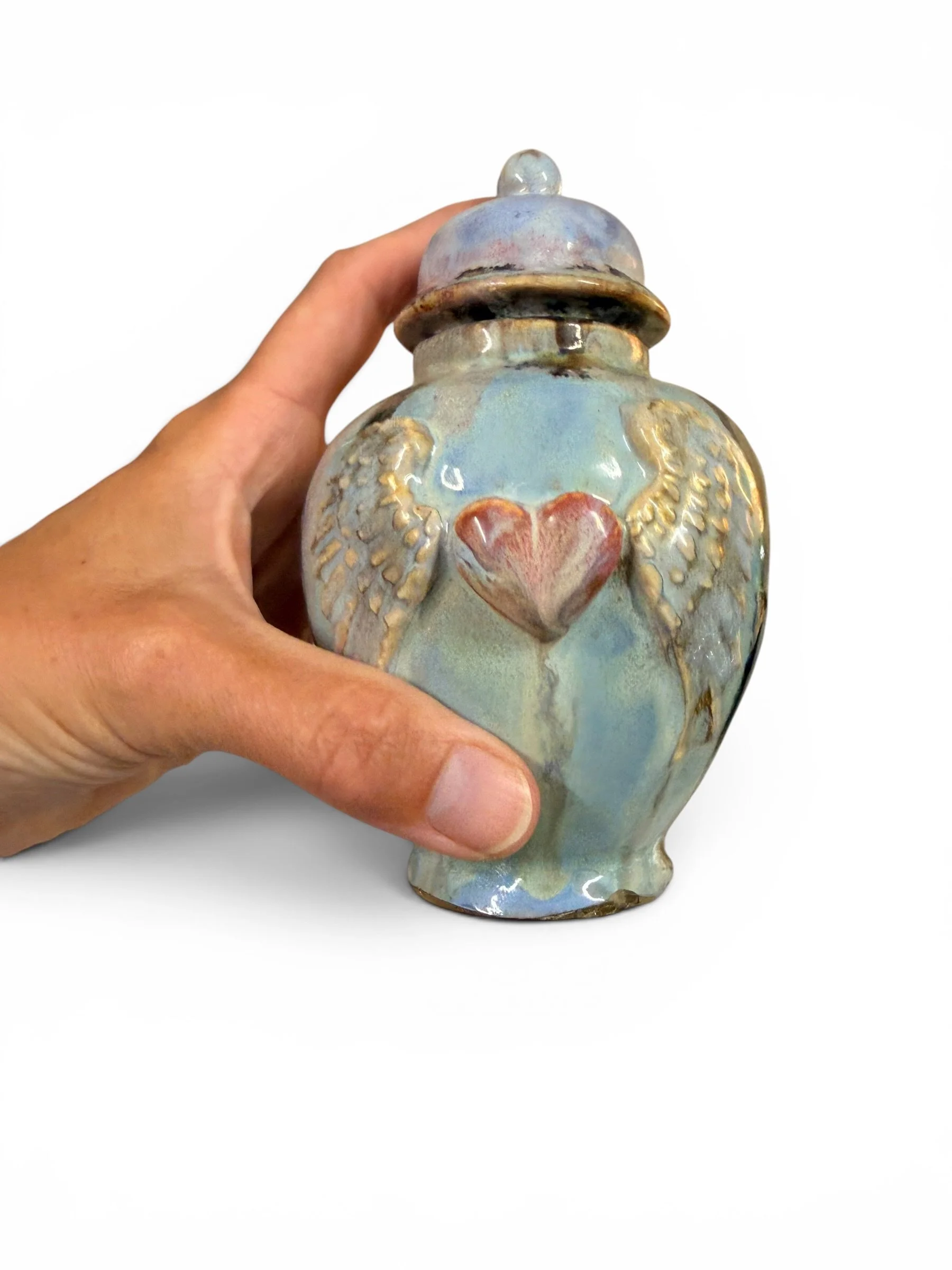 A person holding a ceramic decorative jar with a lid, featuring a heart and bird design, glazed in pastel colors with a shiny finish.