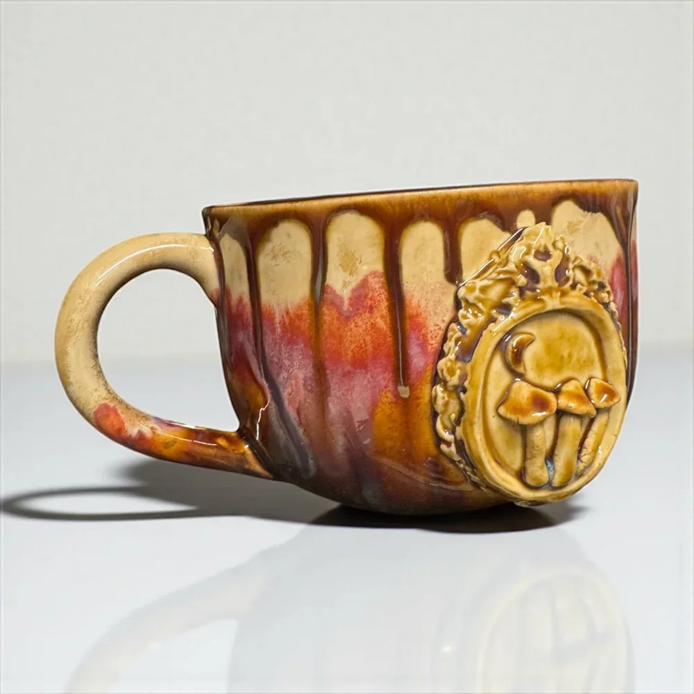 A ceramic mug with a brown, red, and cream glaze, featuring a pumpkin face relief attached to the side.