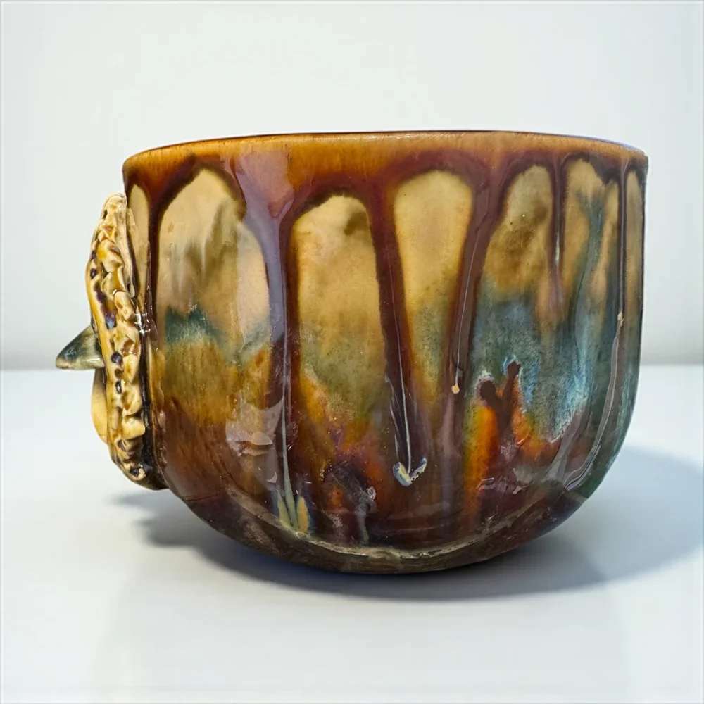 A ceramic bowl with a glossy, multicolored glaze featuring streaks of brown, gold, blue, and green, with a decorative handle shaped like a lion's head on the side.
