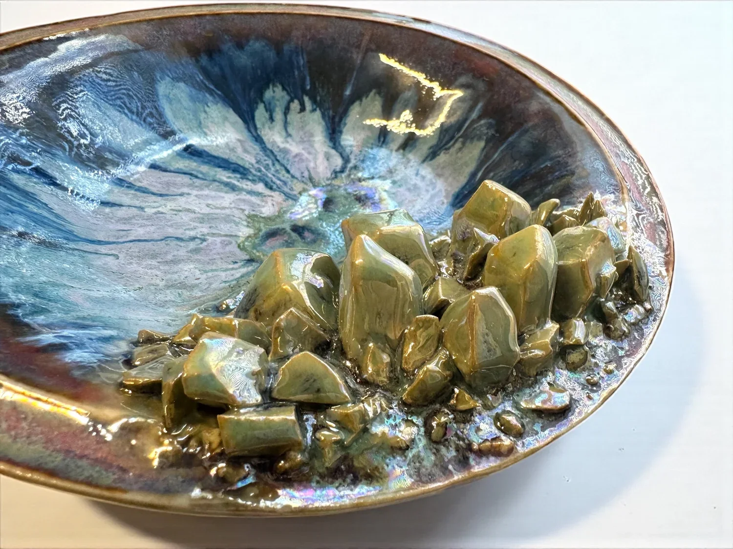 Decorative metal bowl with swirling iridescent blue, green, and purple patterns, filled with irregularly shaped, glossy, greenish-brown stones.