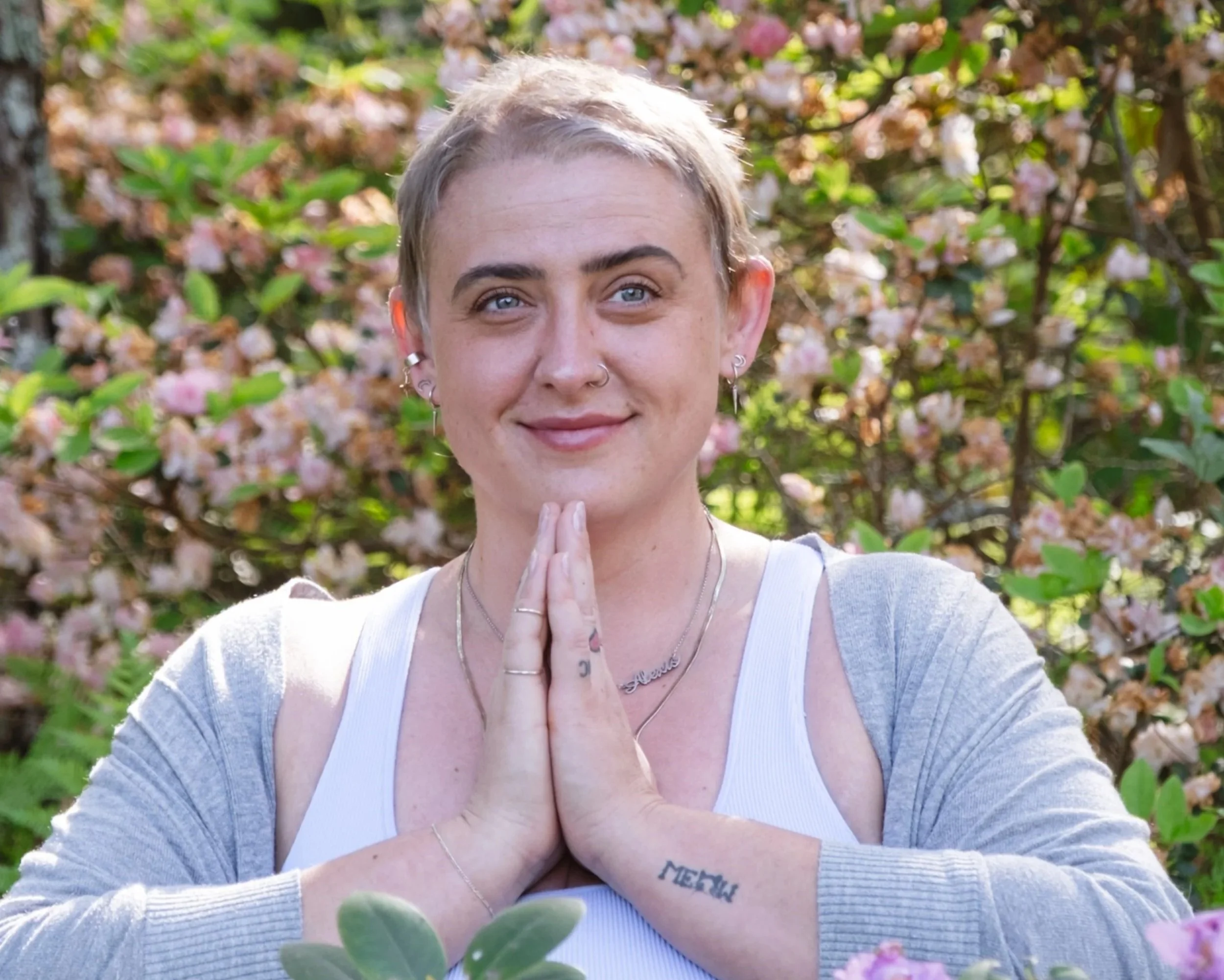 A woman with short blonde hair and multiple earrings standing outdoors with pink flowers and greenery in the background. She is smiling and has her hands pressed together in a prayer or meditation pose.