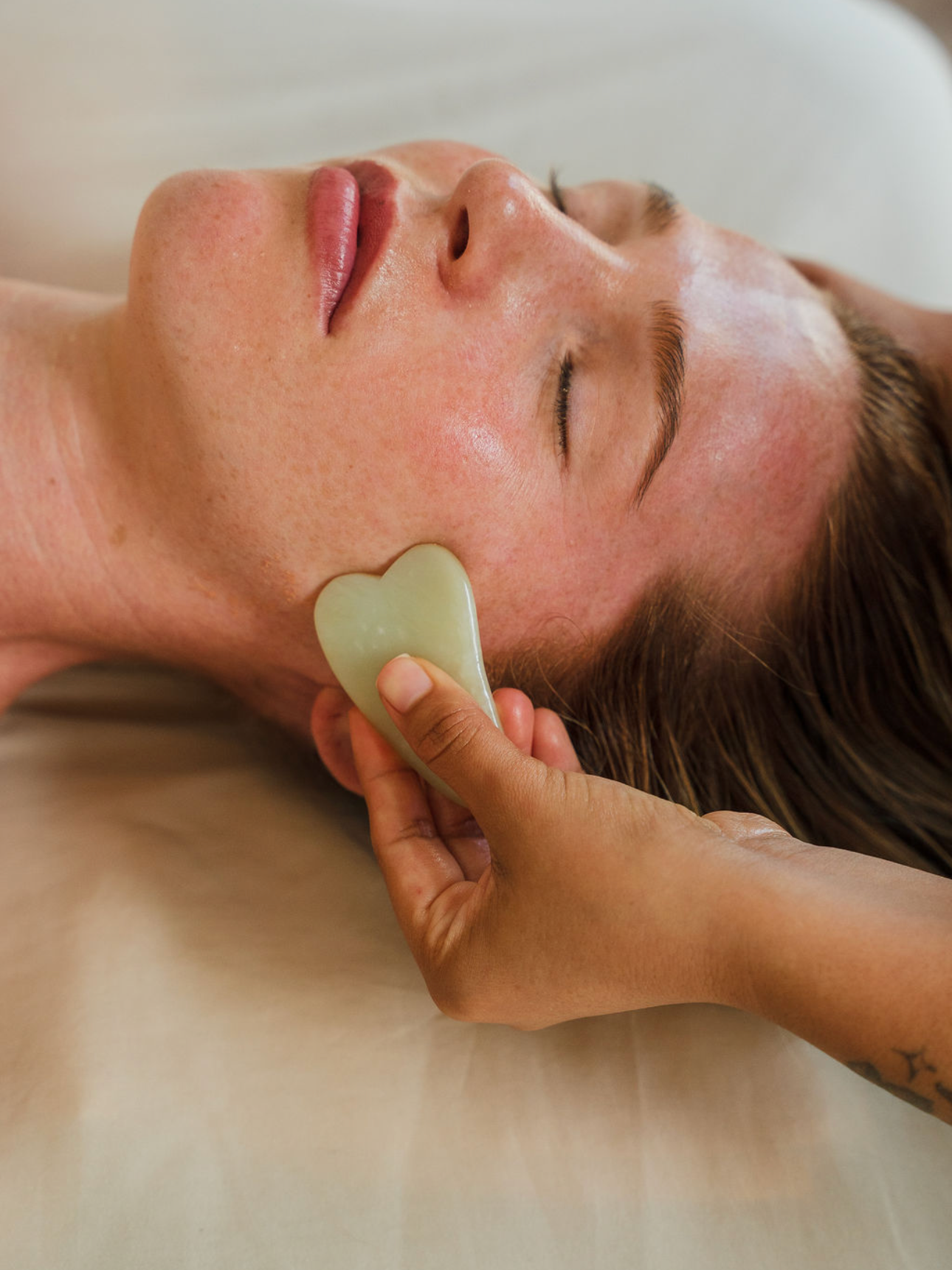 A person receiving a facial treatment with a jade roller on their cheek, lying down with closed eyes.