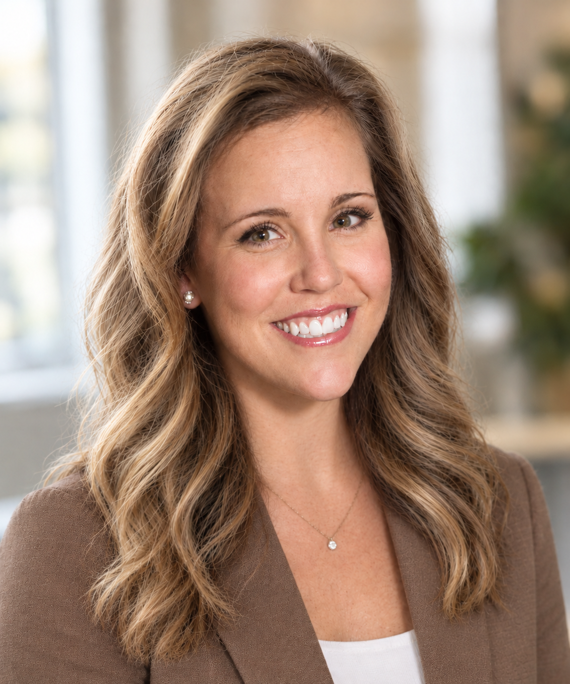 Portrait of a smiling woman with wavy blonde hair, wearing pearl earrings and a necklace, wearing a brown blazer.