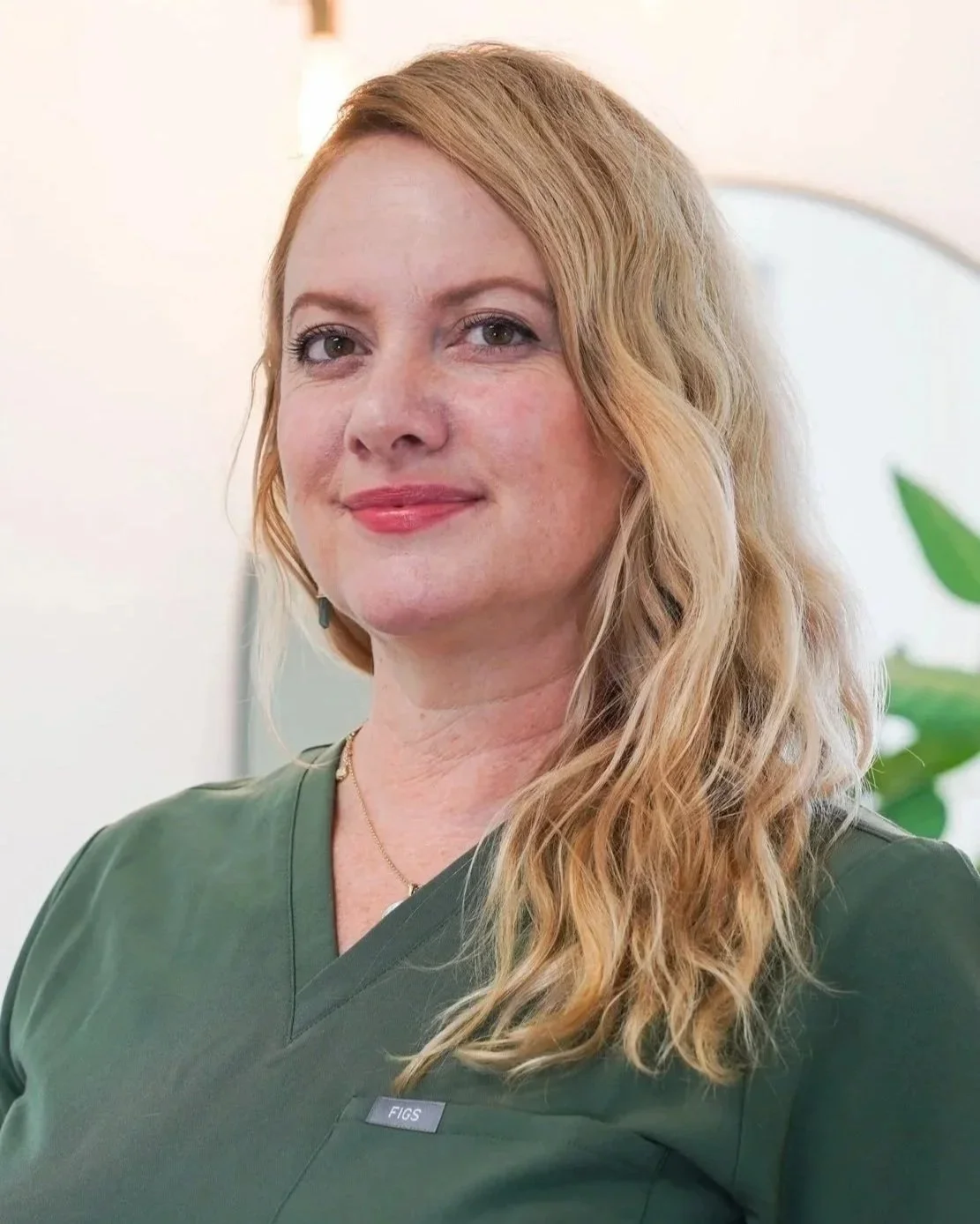 A woman with long wavy blonde hair wearing a green medical uniform, smiling softly at the camera.