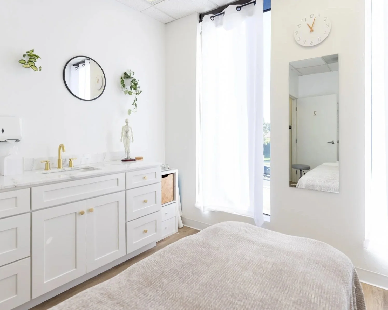 Bright bedroom with white walls, a large window with white curtains, a white dresser with a gold sink faucet, a large mirror, plants, a clock, and a mirror reflections showing a bed and a stool.