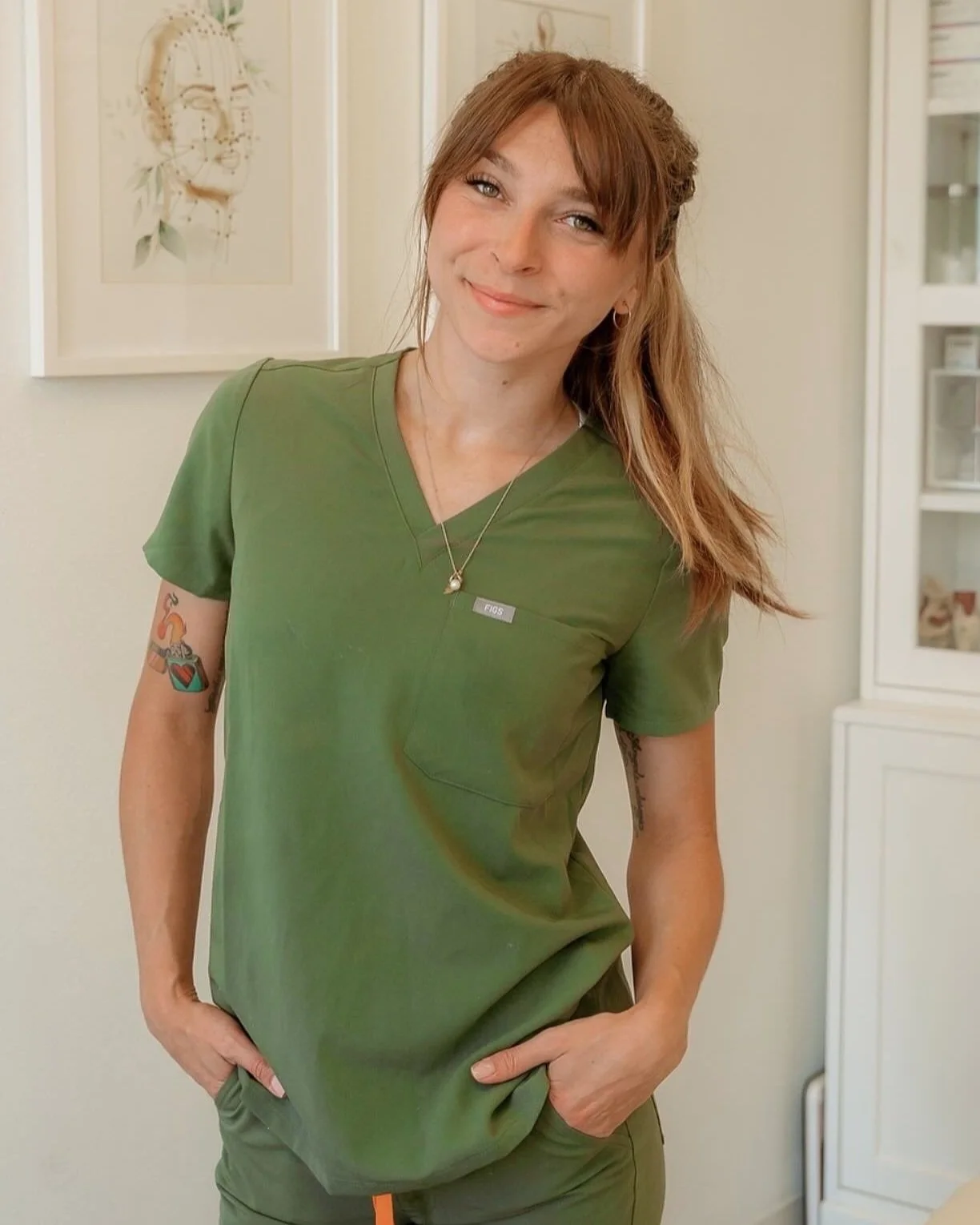 Smiling woman wearing green medical scrubs and a necklace, standing indoors with hands in her pockets.