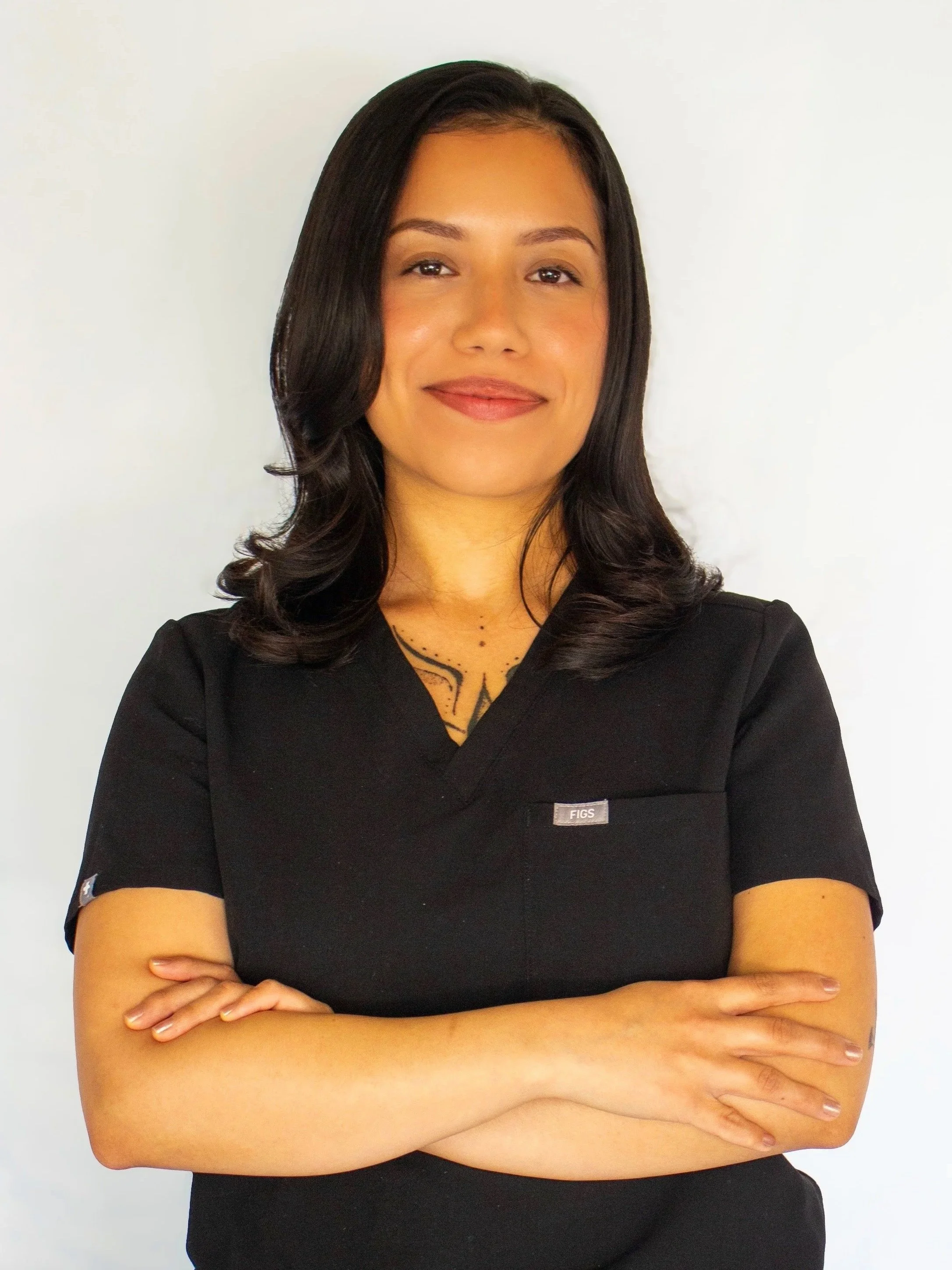 A woman with dark hair in loose waves, wearing a black top with a small logo reading 'FIGS', standing against a white background, with arms crossed and a slight smile.