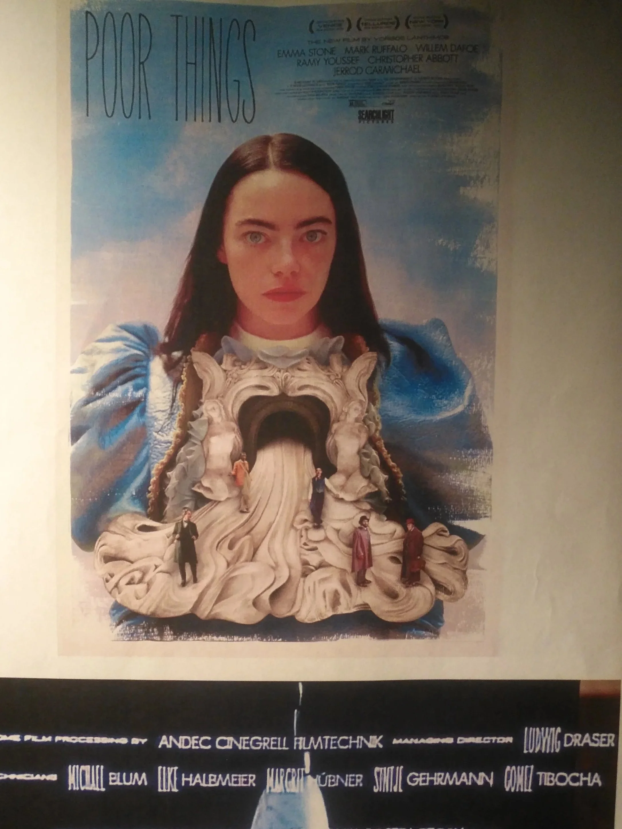 Poster for the film "Poor Things" featuring a woman with dark hair and a serious expression, with a surreal sculptural set piece and miniature figures at the bottom.