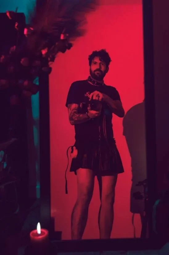Ein Mann steht vor einem Spiegel, hält eine Kamera und trägt einen schwarzen Minikleid mit langen Beinen. Die Szene ist in rotem Licht getaucht.