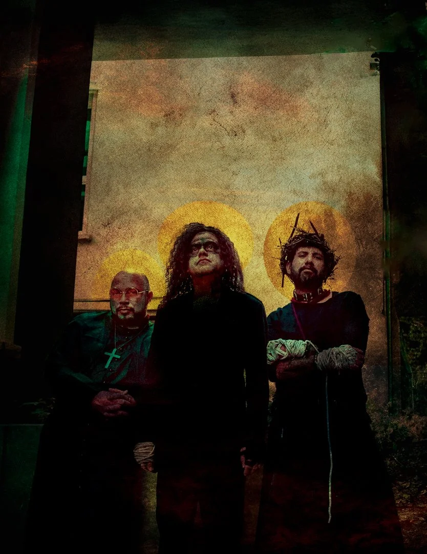 Three individuals standing together in front of a backdrop with three golden halos, one behind each person. The central figure has curly hair and wears dark clothing, looking upward. The person on the left has a shaved head, glasses, a beard, and a c