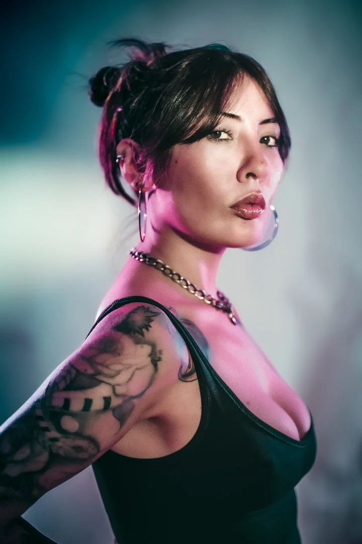 A woman with dark hair and tattoos is sitting in a corner of a concrete structure with curved blue walls, posing with her arm reaching upward and her head tilted back, casting shadows on the ground.