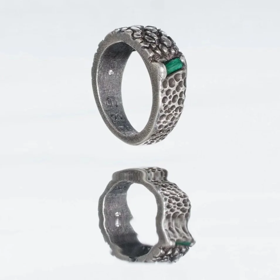 Ein silberfarbener Ring mit einer strukturierten Oberfläche, einem grünen Akzent und einer organischen Form, der auf einer reflektierenden Oberfläche liegt.