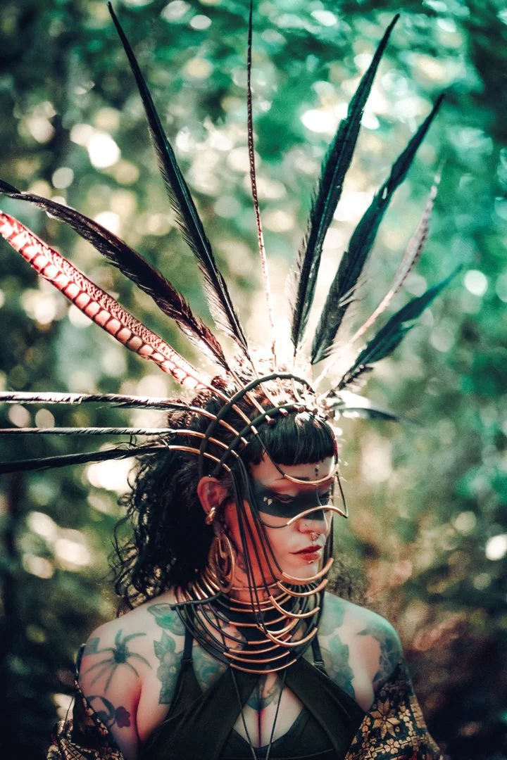 Frau mit schwarzem Make-up, Tattoos, und traditionellem Kopfschmuck aus Federn und metallischen Ringen in einem Wald.