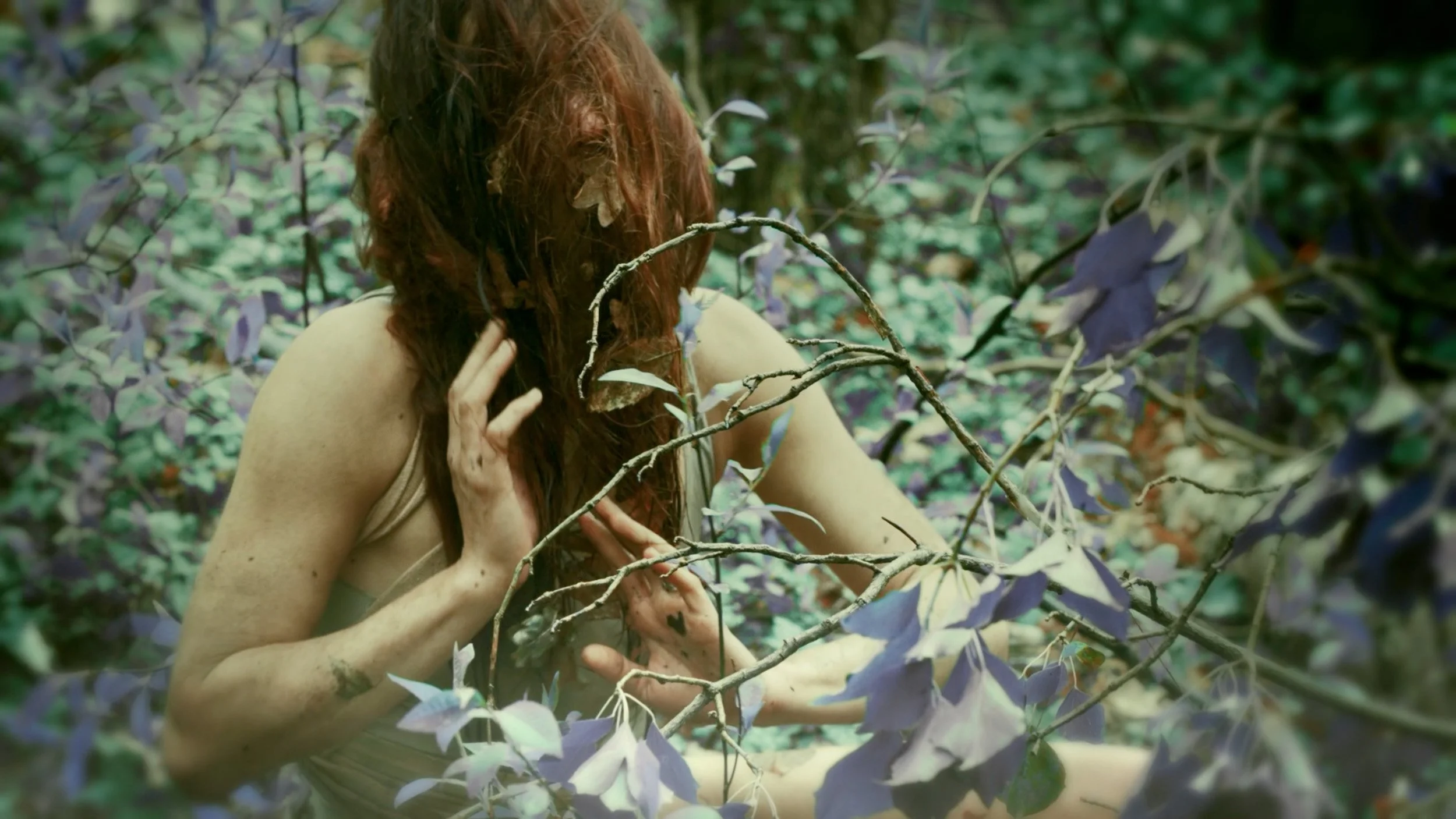 Eine Frau mit roten Haaren in einem Wald, die sich umarmt und hinter Zweigen versteckt.