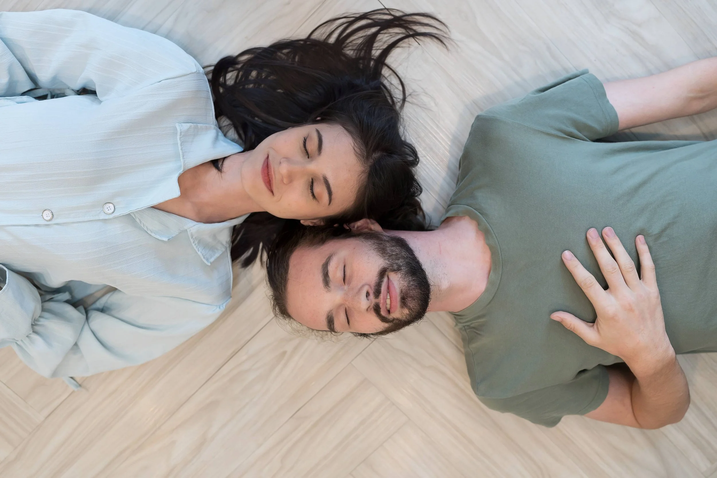 White man and woman lay next to one another, heads together, faces toward camera, and eyes closed during a ketamine couples therapy session. Man holds hand over his heart.