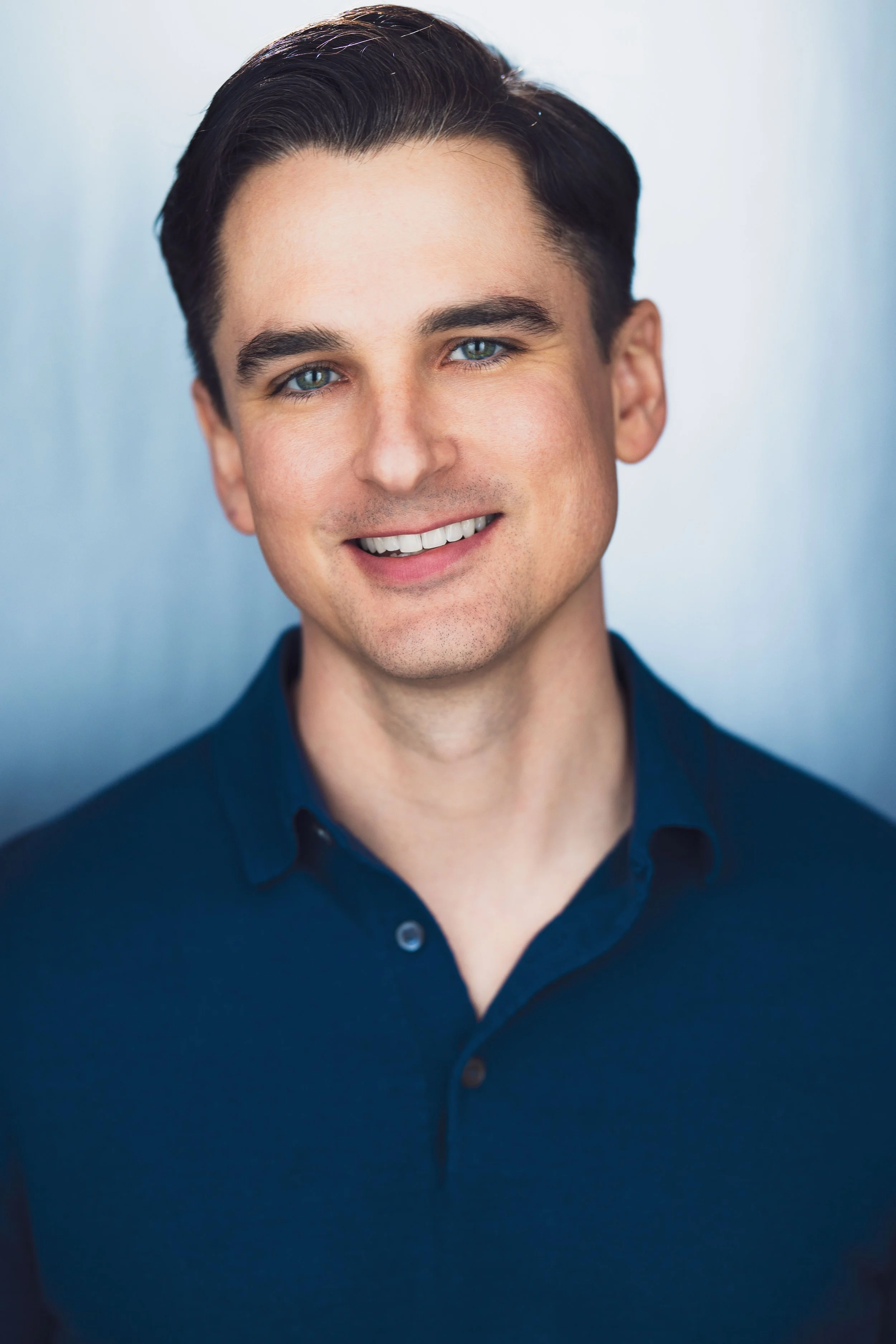 Close-up of a young man with dark hair, blue eyes, and a bright smile, wearing a dark blue button-up shirt.