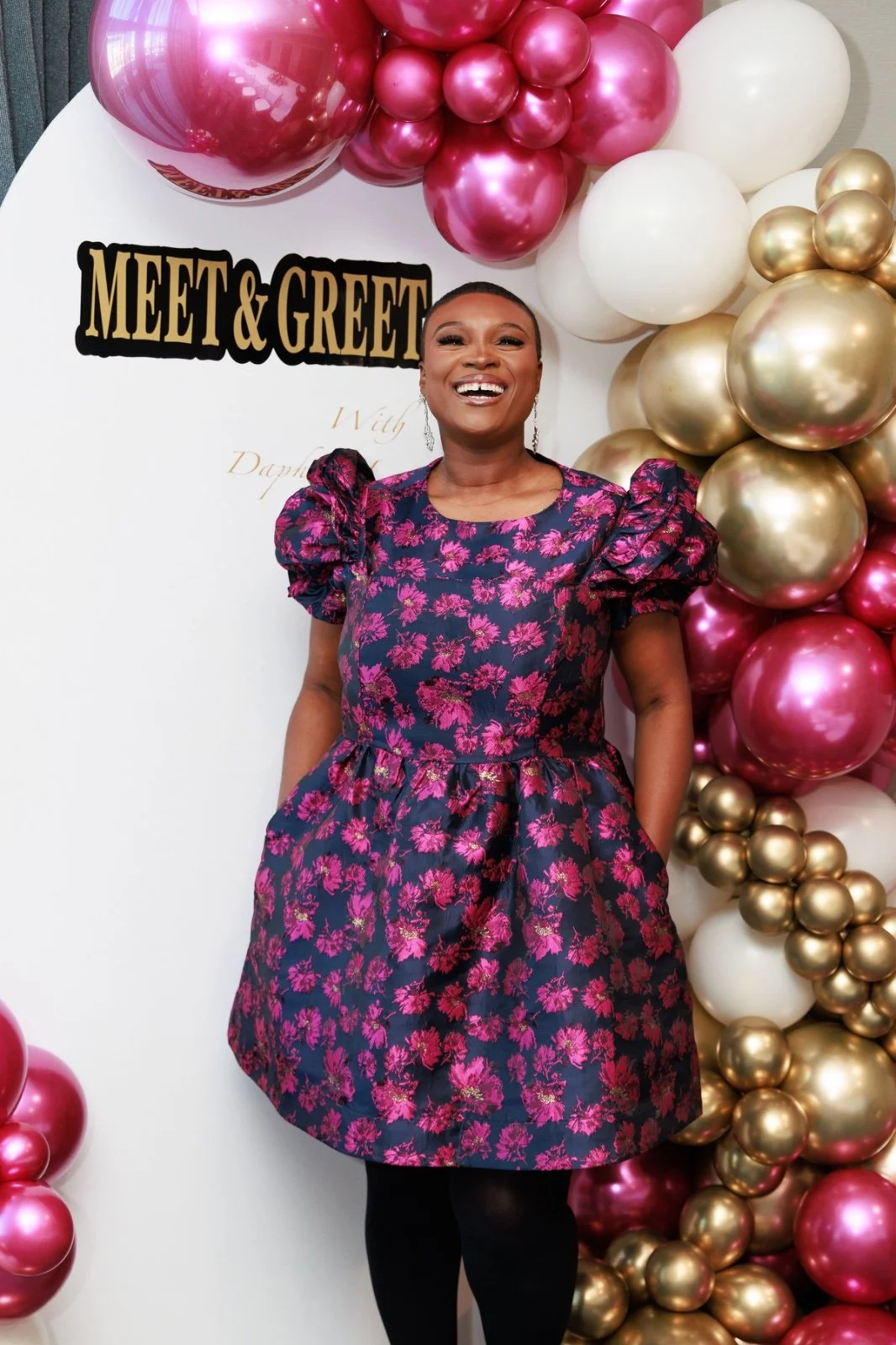 A woman in a navy blue and pink floral dress smiling at a meet and greet event, standing in front of a white backdrop decorated with pink, white, and gold balloons.