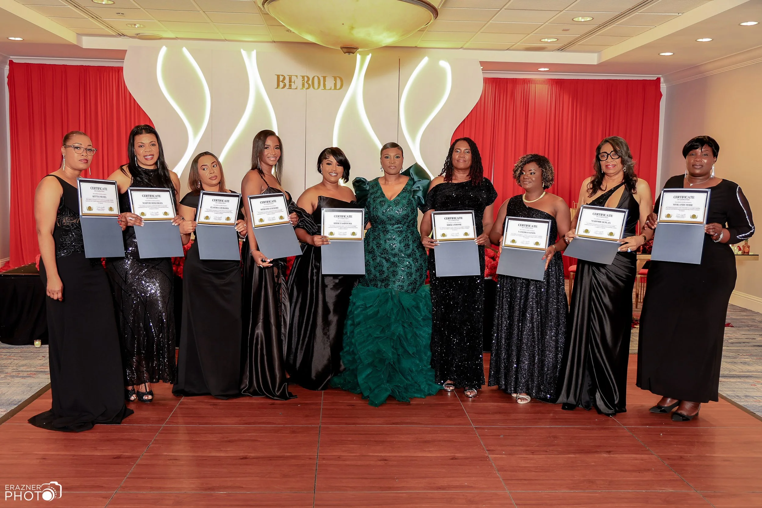Group of ten women dressed in formal gowns, standing indoors, holding certificates, with a backdrop that reads 'BE BOLD.'