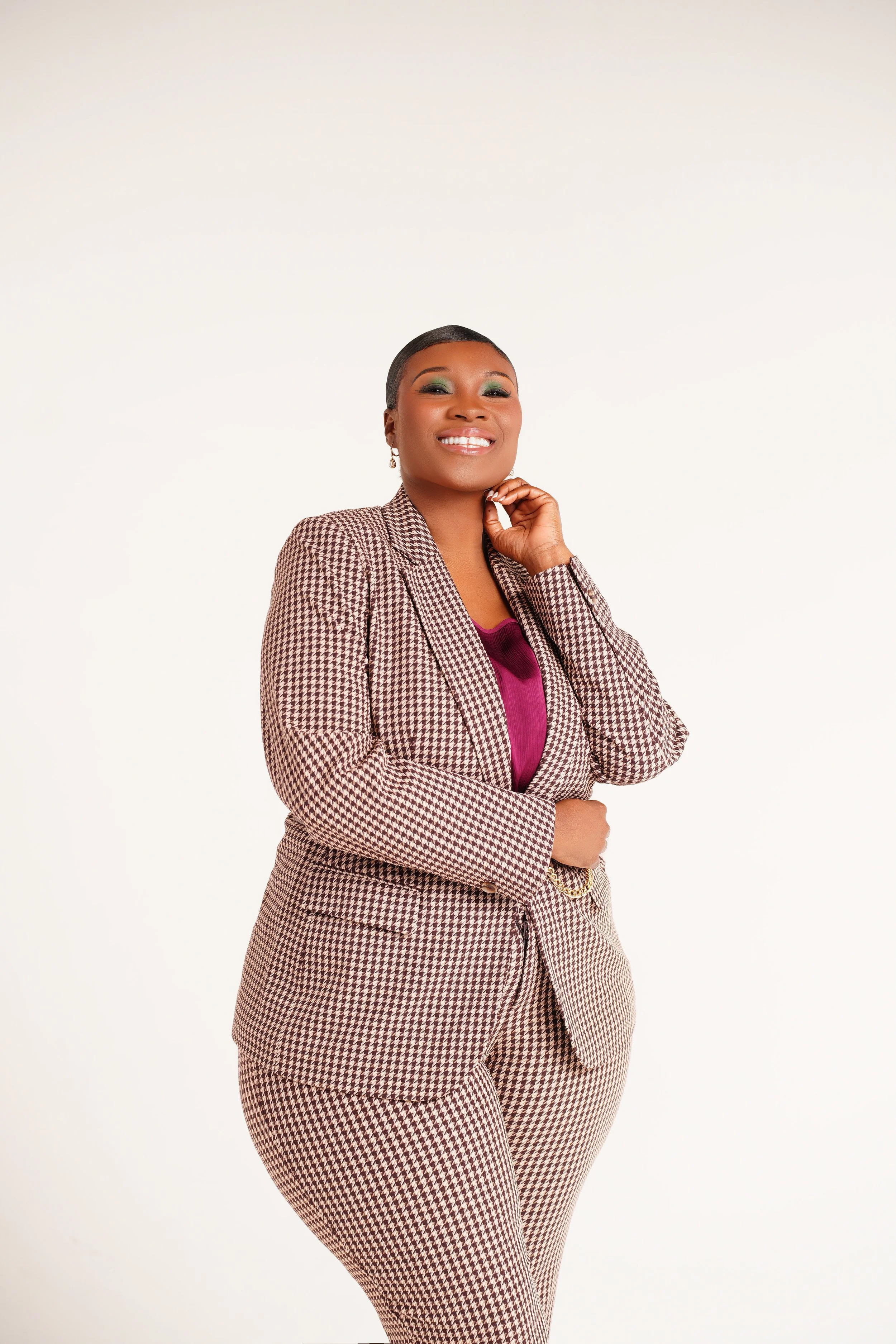 A smiling woman in a houndstooth suit with a purple top, standing with one hand near her face against a plain white background.