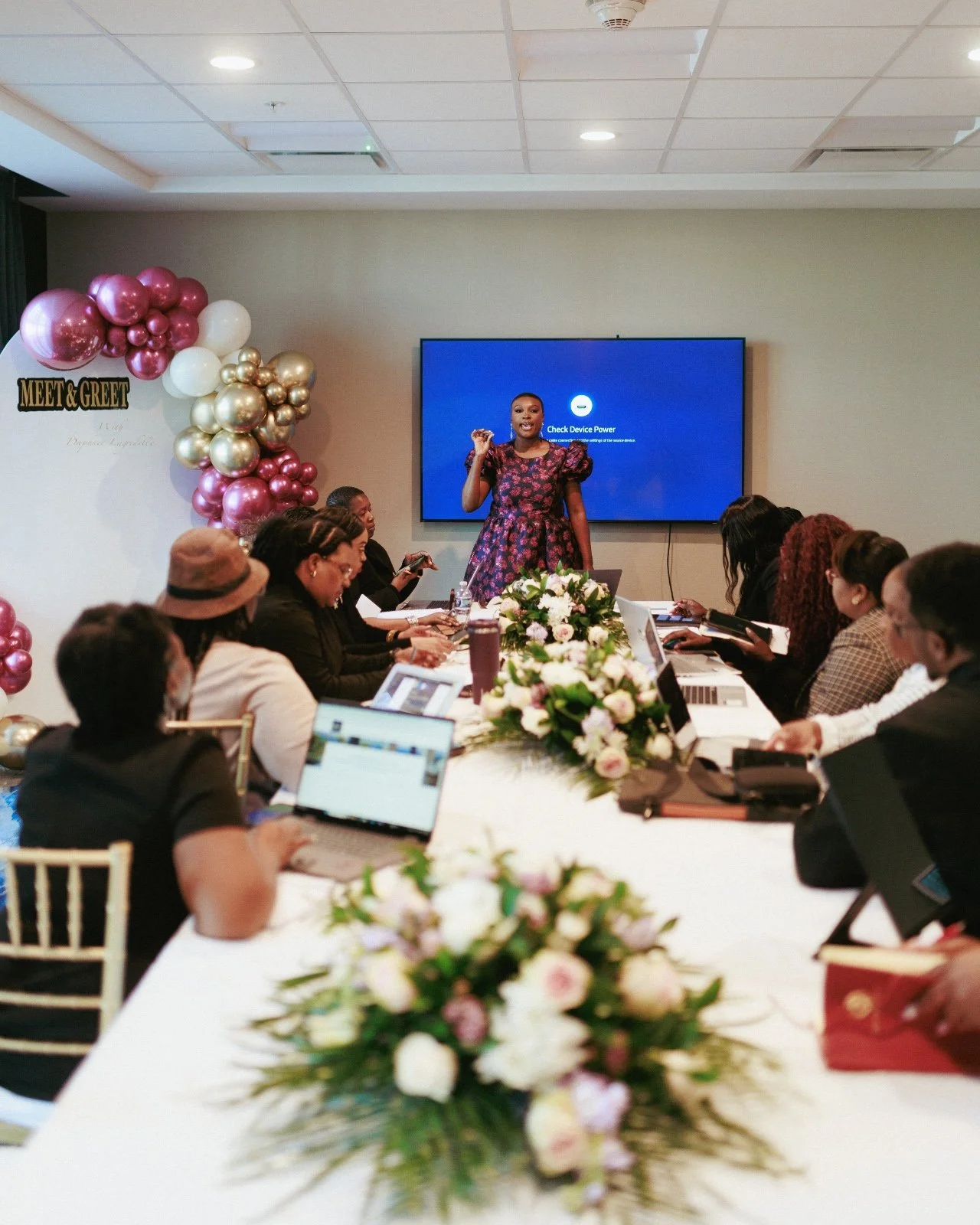 A woman is giving a presentation at a meet and greet event with balloons and flowers decorating the room. Attendees are seated around a table with laptops, listening attentively.