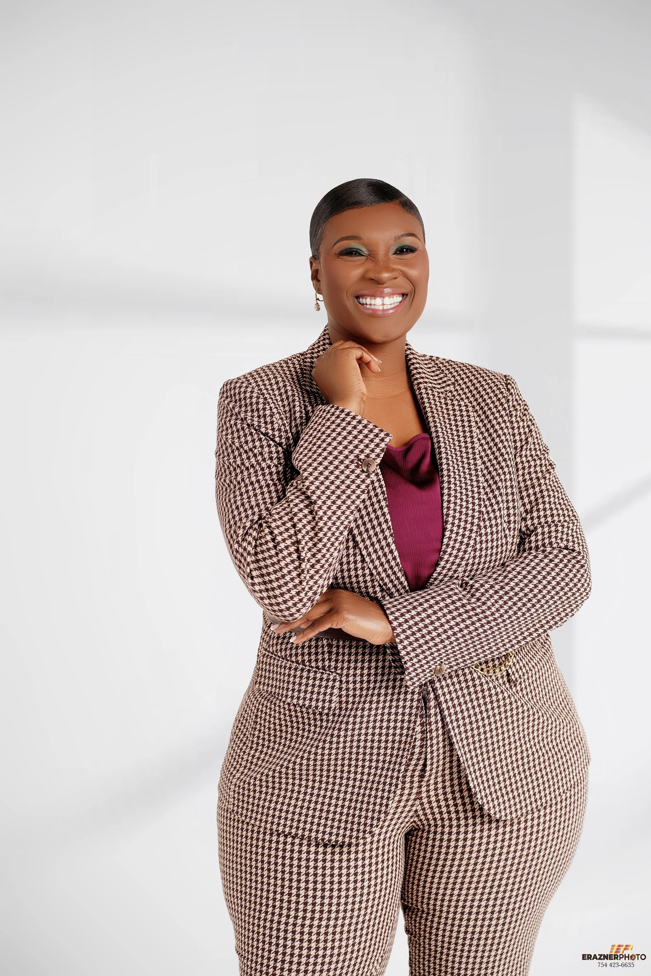 A confident woman smiling, with short dark hair, wearing a brown and beige houndstooth suit and a maroon blouse, standing against a bright white background.