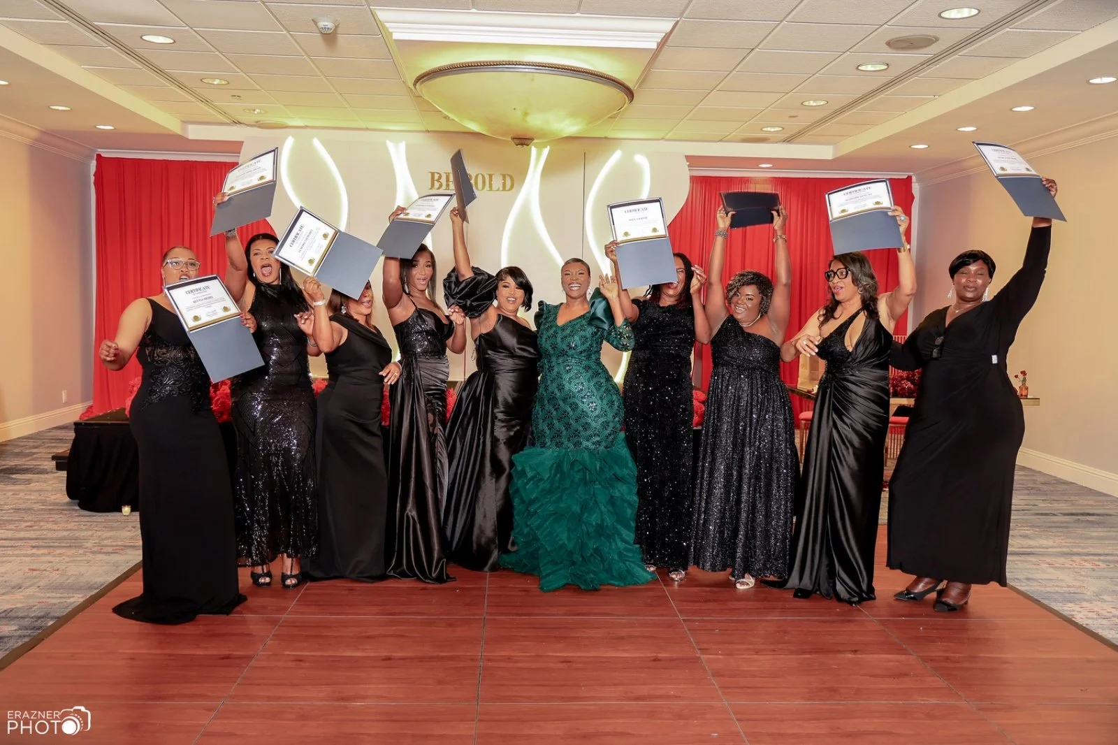 A group of women in formal black dresses celebrating and holding up certificates at an awards ceremony in a decorated banquet hall with a red curtain backdrop.