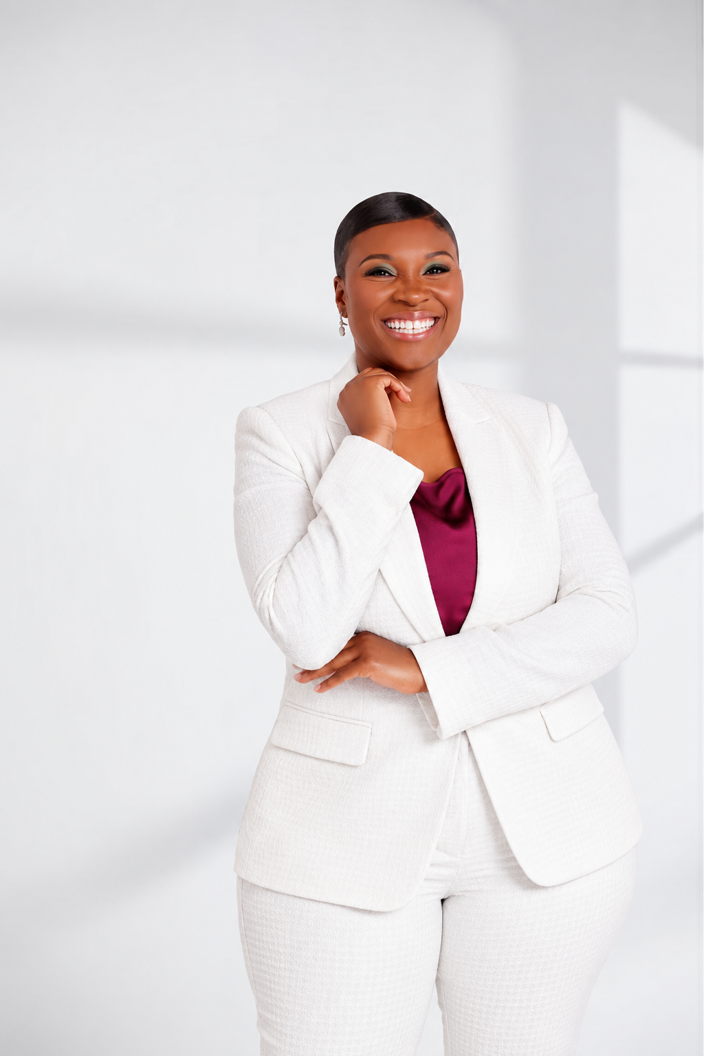 A confident African American woman with short black hair smiling, wearing a white suit and a maroon blouse, standing with one hand near her chin and the other crossed over her chest in a bright, minimal office setting.