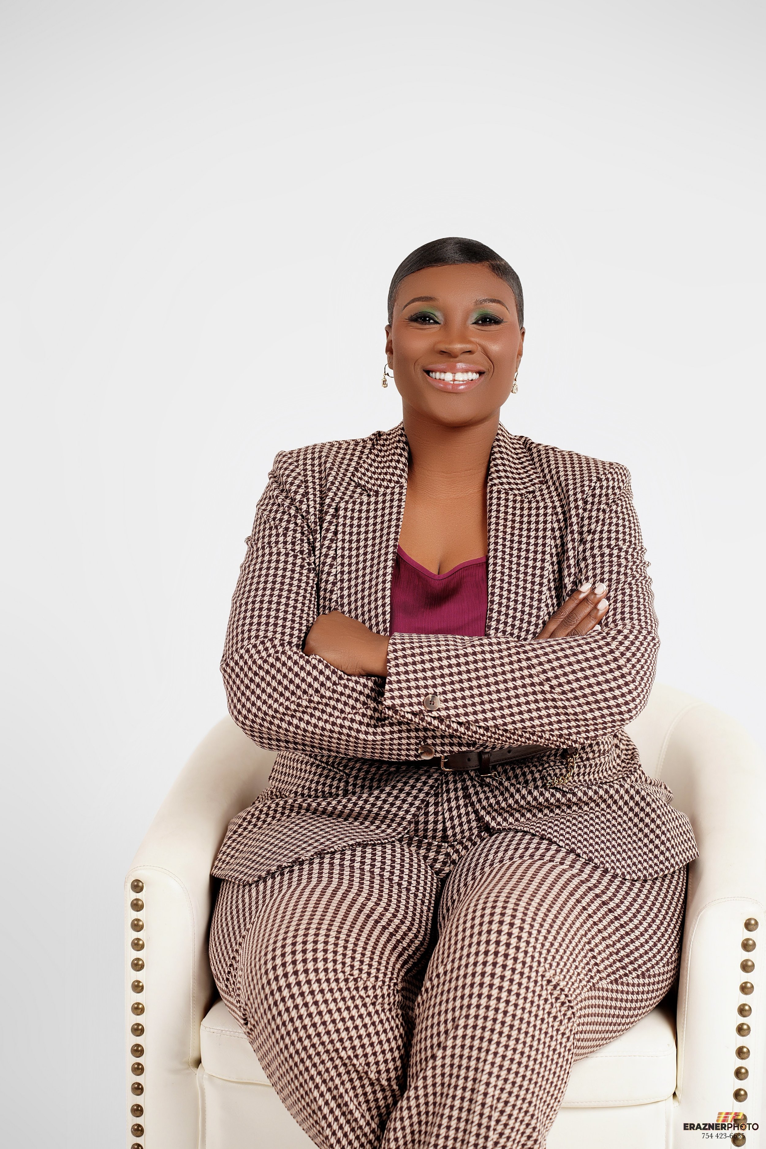 A woman sitting in a white chair, smiling, with arms crossed, wearing a brown houndstooth suit and a maroon top, neutral background.