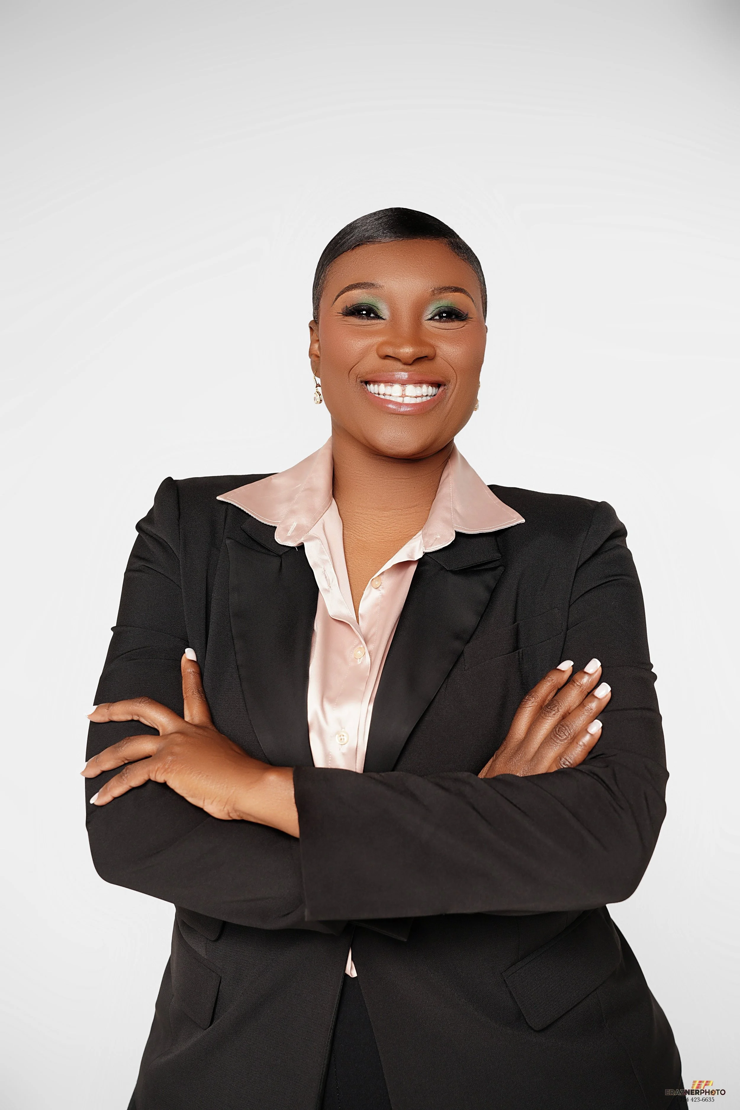 Professional woman smiling with arms crossed, wearing a black blazer and light pink satin blouse, against a white background.