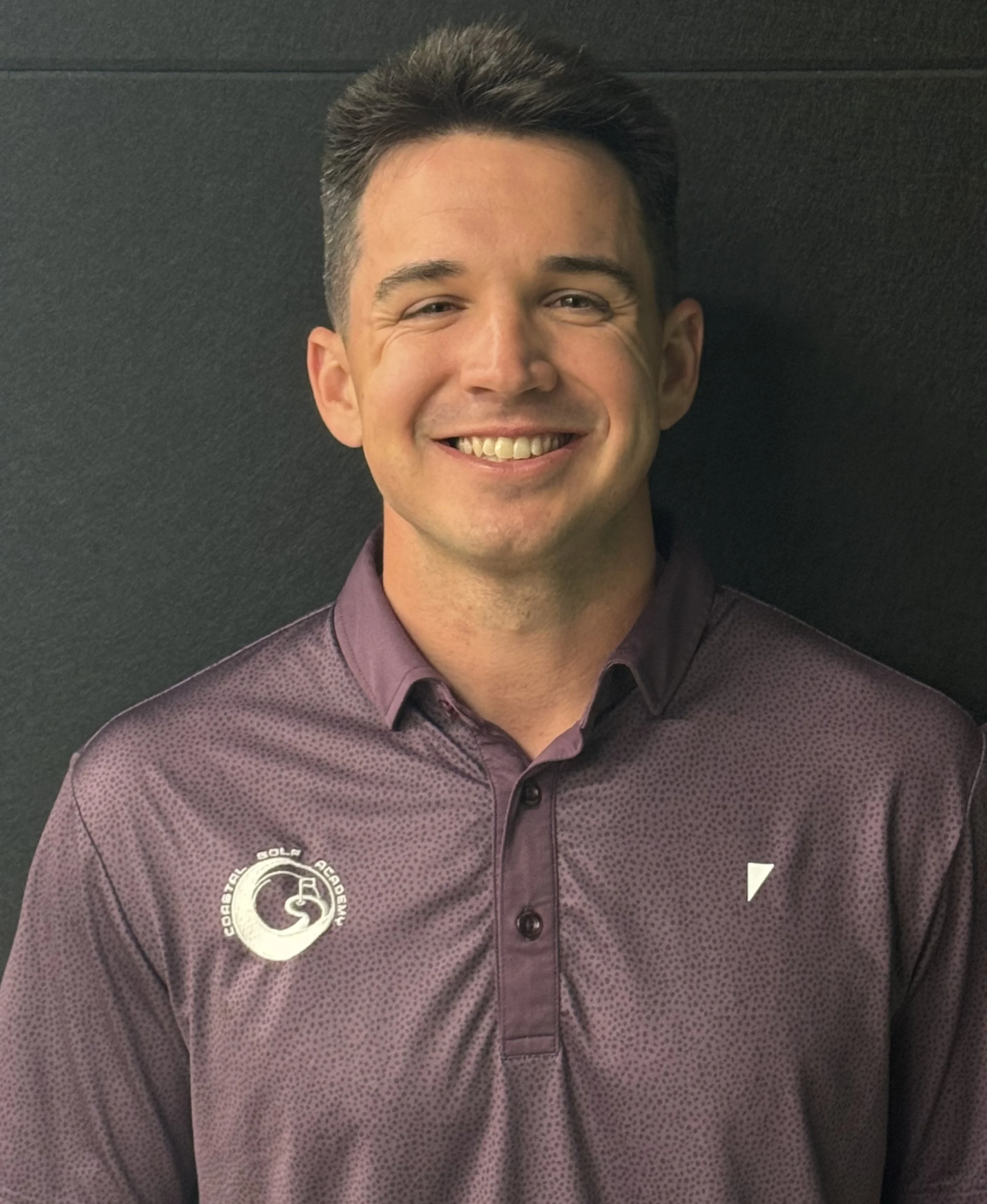 Lawson short dark hair, smiling, wearing a maroon collared shirt with a logo on the chest, sitting against a dark background.