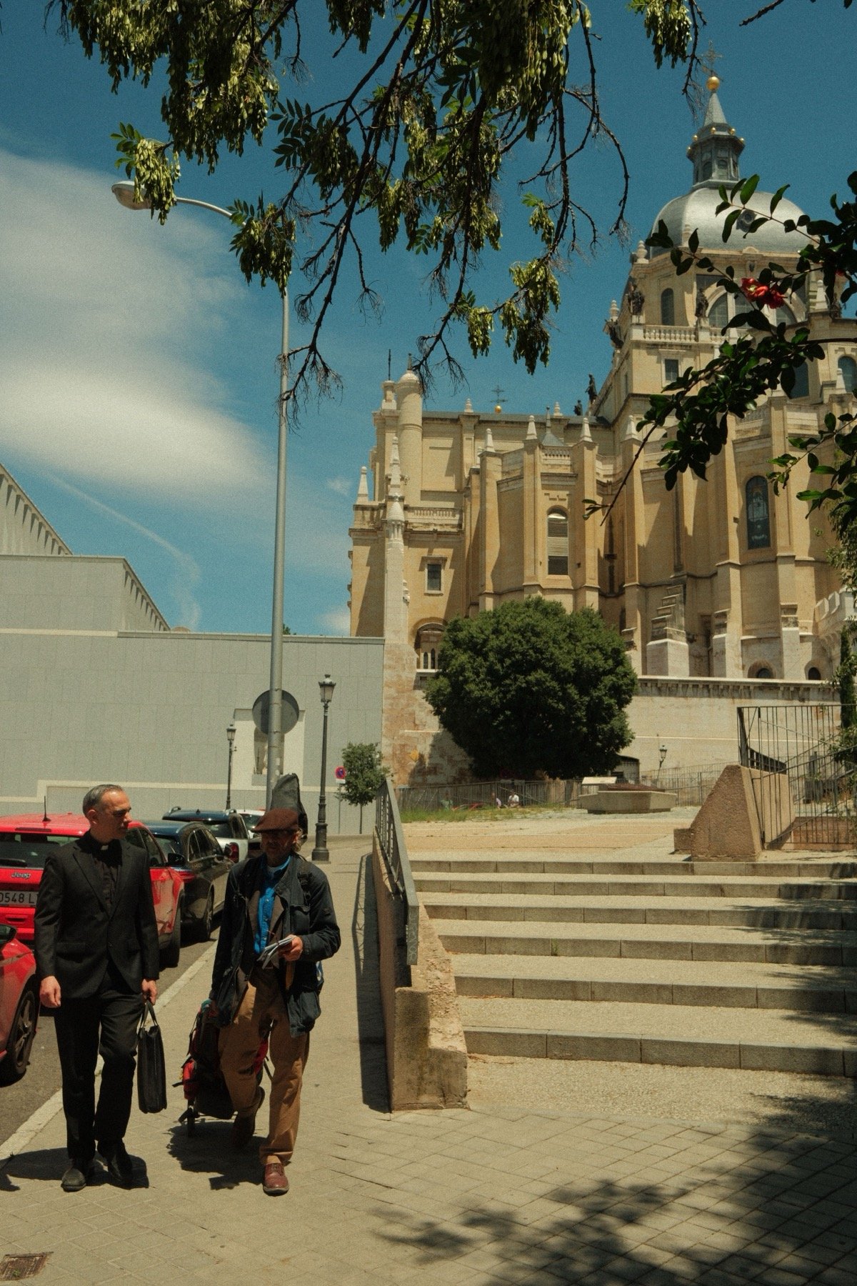 Two men walking near the steps of Santa Maria Cathedral in Madrid under blue skies